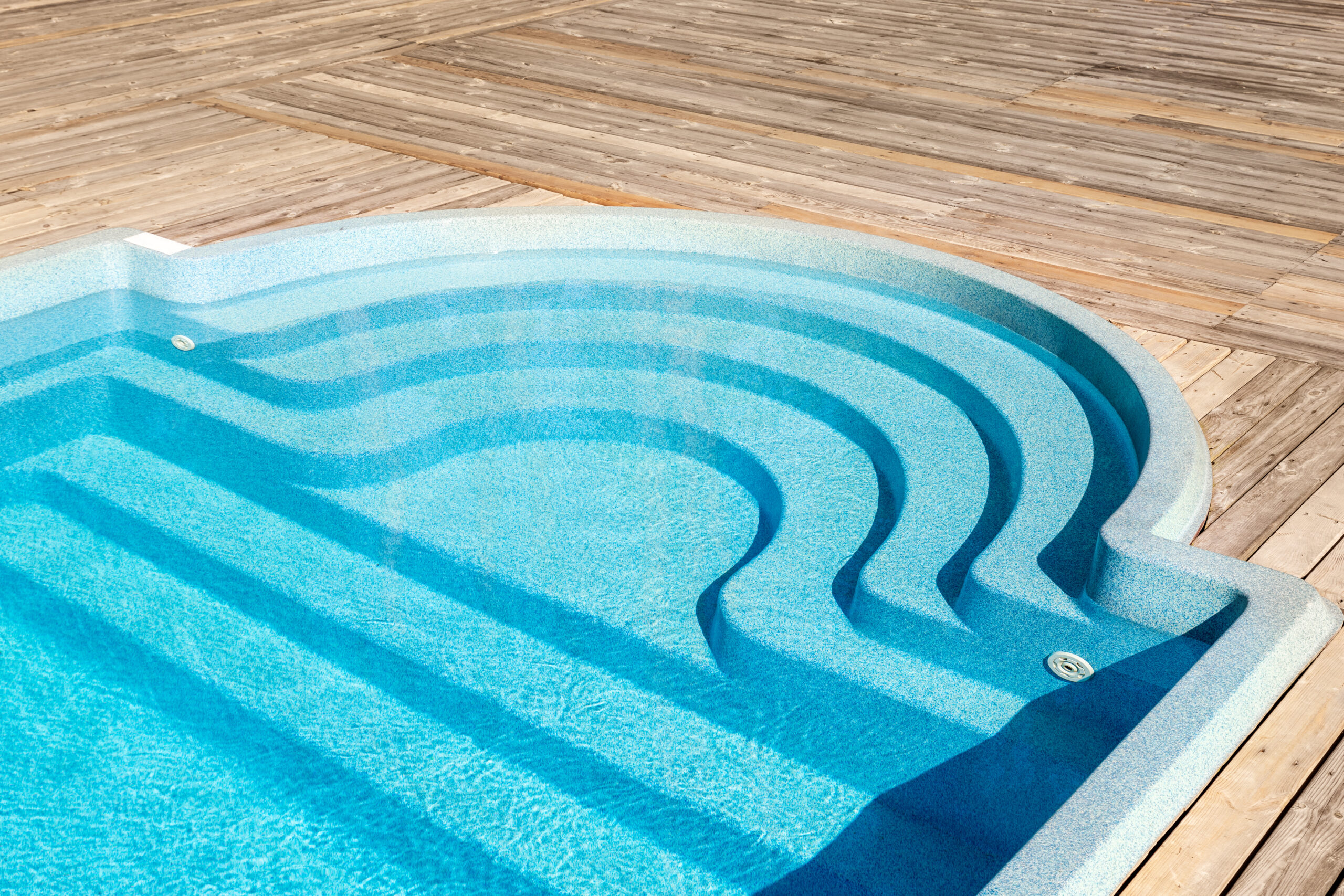 New modern fiberglass plastic swimming pool entrance step with clean fresh refreshing blue water on bright hot summer day at yard or resort hotel spa area. Wooden flooring deck of teak or larch board.