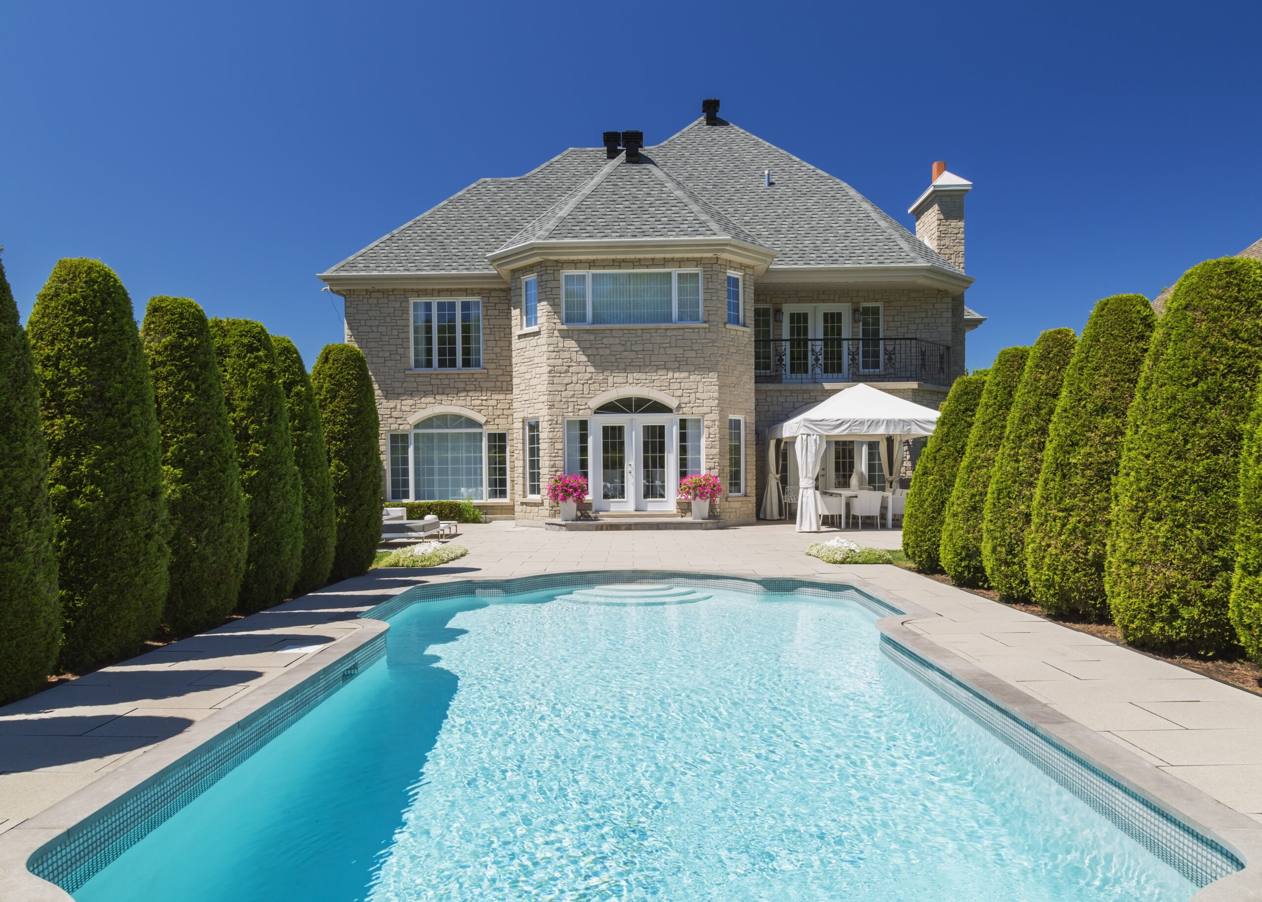 Outdoor swimming pool bordered by rows of cedar trees (Thuja occidentallis) at rear of luxurious house with beige stone