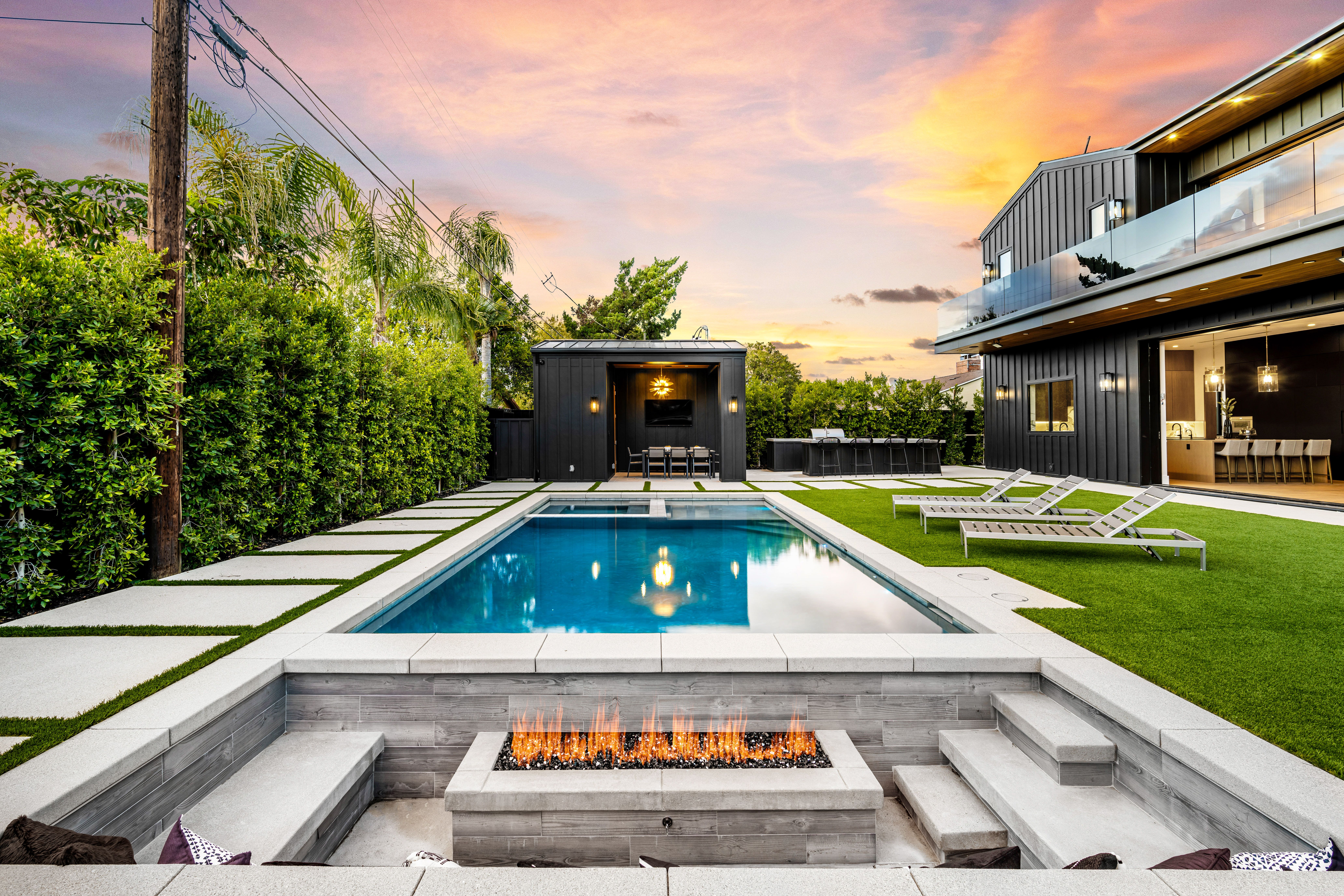 Modern backyard with a swimming pool, fire pit, and lounge chairs at sunset, featuring a contemporary house with large windows and manicured garden.