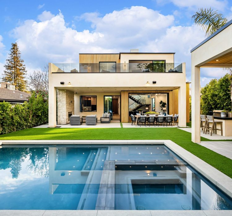 Modern luxury house with a swimming pool, outdoor seating area, and lush greenery under a bright blue sky.